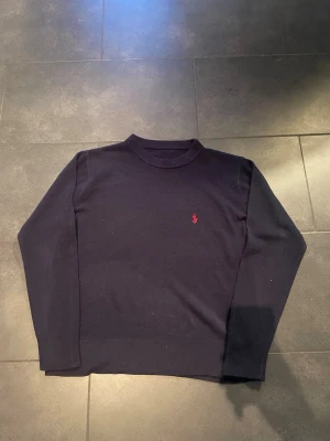 Mörkblå tröja från Ralph Lauren - Klassisk mörkblå tröja från Ralph Lauren med rund hals och diskret röd logga broderad på bröstet. Tröjan har lång ärm och ribbade muddar vid ärmslut och nederkant. Perfekt till lager på lager under kyliga dagar.
