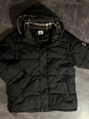 Svart pufferjacka från Burberry - Säljer en svart pufferjacka från Burberry med klassiskt rutigt foder i huvan. Jackan har dragkedja, knappar och en patch med Burberry London England på ärmen. Perfekt för kalla dagar och riktigt snygg streetstil.