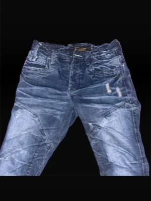 Blå Fishbone jeans med snörning - Säljer ett par blå jeans från Fishbone med coola detaljer som snörning vid benslutet och slitningar framtill. Jeansen har flera fickor, markerade sömmar och en relaxed passform. Perfekta för dig som gillar streetwear och vill sticka ut lite extra.