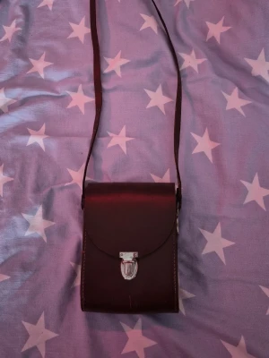 Mörkbrun axelväska i skinn - Säljer en stilren mörkbrun axelväska i skinn med enkel design och silverfärgat spänne framtill. Väskan har ett långt axelband och röd insida, perfekt för dig som vill ha en liten och smidig väska till mobilen och småprylar.