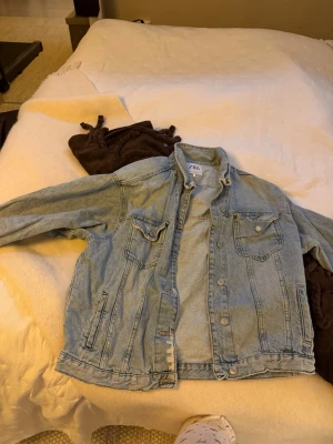 Ljusblå jeansjacka från Zara - Säljer en ljusblå jeansjacka från Zara med klassisk krage, bröstfickor med knappar och slitna detaljer på ena ärmen. Jackan har långa ärmar och silverfärgade knappar framtill. Perfekt för dig som gillar en chill och trendig look.