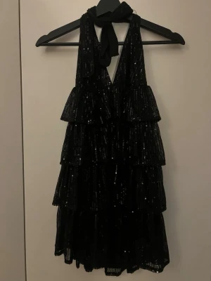 Svart glittrig volangklänning - Svart glittrig volangklänning med öppen rygg och knuten i nacken. Smidigt att kunna knyta den hur kort/lång du vill ha den. Aldrig använd