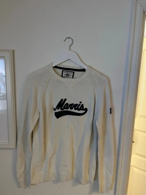 Vit stickad tröja från Morris - Vit stickad tröja från Morris med svart logga framtill och gråa armbågslappar. Tröjan har rund halsringning, ribbade muddar och lång ärm. Perfekt för dig som gillar klassisk stil med en twist.