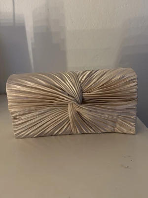 Champagnefärgad kuvertväska i satin - Snygg kuvertväska i champagnefärgad satin med veckad framsida och twistdetalj. Väskan har en tunn silverfärgad kedja som axelrem och stängs med magnetlås. Perfekt storlek för det viktigaste och ger en elegant touch till din outfit.