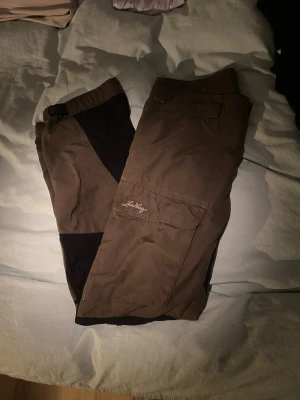 Bruna friluftsbyxor från Lundhags strl 42 - Säljer ett par bruna friluftsbyxor från Lundhags i storlek 42. Byxorna har svarta förstärkningar på knäna, stora fickor på benen och är tillverkade i slitstarkt bomullsmaterial. Perfekta för uteliv och äventyr.