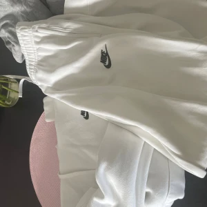 Vita mjukisbyxor från Nike - Säljer ett par vita mjukisbyxor från Nike med svart logga på låret. Byxorna har resår i midjan och är tillverkade i mjukt material som passar perfekt för chill eller träning. Klassisk och enkel design med fickor på sidorna. Set byxor och tröja 