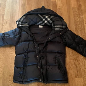 Burberry Jacka Svart - Snygg svart dunjacka från Burberry med klassiskt rutigt foder i huvan. Jackan har dragkedja och knappar framtill, två fickor med dragkedja och en patch med Burberry London England på ärmen. Perfekt för kalla dagar och riktigt stilren look.