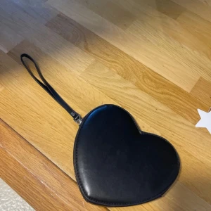 Svart hjärtformad plånbok med dragkedja - Snygg svart plånbok i hjärtform med handledsrem och dragkedja runt hela kanten. Perfekt för att bära småsaker eller kort. Stilren design som sticker ut och är lätt att ta med sig överallt.