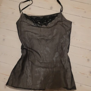 Svart mesh linnetopp med pärlbroderi - Säljer en svart mesh linnetopp med tunna justerbara axelband och snyggt pärlbroderi framtill vid urringningen. Toppen är lätt transparent och har en figurnära passform, perfekt för att skapa en cool och trendig look.