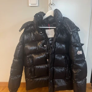 Svart Moncler pufferjacka med huva - Säljer en svart pufferjacka från Moncler med glansig finish, stor huva och klassisk logga på ärmen. Jackan har dragkedja, knappar och flera fickor. Insidan har ett coolt serietidningsmärke. Perfekt för kalla dagar när du vill hålla stilen.