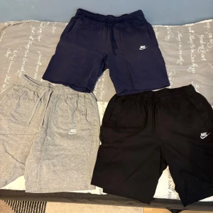 Nike shorts i tre färger med snörning - Tre par Nike shorts i svart, marinblått och ljusgrått. Alla har elastisk midja med snörning, sidofickor och klassisk Nike-logga broderad på vänster ben. Perfekta för sommaren och chill dagar. Mjukt bomullsmaterial för maximal komfort.