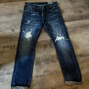 Jack & Jones mörkblå jeans med slitningar - Säljer ett par mörkblå jeans från Jack & Jones med coola slitningar och trasiga detaljer på framsidan. Jeansen har klassisk femficksdesign, raka ben och kontrastsömmar. Materialet är robust denim i bomull. Perfekt för dig som gillar en edgy streetstil. Skulle säga storlek L men står 33/34 Tall 
