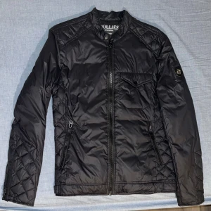 Svart lättviktsdunjacka Hollies S - Svart lättviktsdunjacka från Hollies med quiltade detaljer på axlar och ärmar. Jackan har dragkedja framtill, bröstficka med knapp och två snedställda fickor med dragkedja. Snygg badge på ena ärmen och stilren, smal passform.