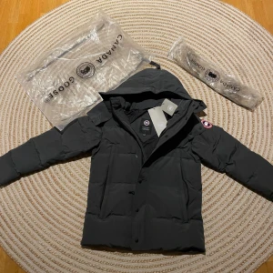 Svart dunjacka från Canada Goose M - Säljer en mörk grå dunjacka från Canada Goose i storlek M. Jackan har en avtagbar huva med naturfärgad pälskant, klassisk logga på ärmen och flera praktiska fickor. Perfekt för kalla vinterdagar och riktigt snygg streetstil. Den är helt ny