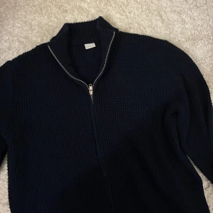 Mörkblå stickad kofta med dragkedja - Mörkblå stickad zip tröja med grov struktur och hel dragkedja framtill. Hög krage och långa ärmar gör den perfekt för kyligare dagar. Enkel och stilren design som passar till det mesta.