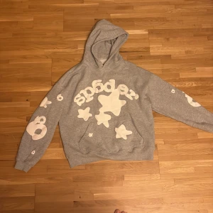 Grå Sp5der hoodie med vita tryck - Säljer en grå hoodie från Sp5der med stora vita stjärntryck och logga framtill samt tryck på ärmarna. Tröjan har huva och känguruficka, tillverkad i mjuk bomull. Perfekt för dig som gillar streetwear och vill sticka ut.