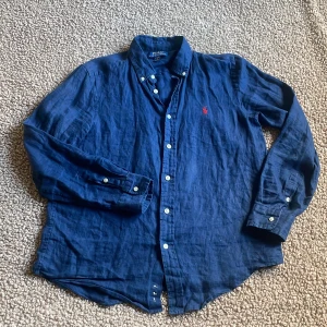 Blå linneskjorta från Polo Ralph Lauren - Snygg marinblå skjorta i linne från Polo Ralph Lauren med klassisk button-down krage och röd broderad logga på bröstet. Skjortan har långa ärmar och vita knappar framtill. Passar dig mellan 160~170cm. Skriv gärna vid några fundering kring skjortan!                MHV SWISSWEAR🇨🇭🍾🏔️