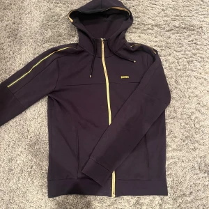 Svart hoodie från BOSS med dragkedja - Svart hoodie från BOSS med gul dragkedja och gul logotyp på bröstet. Tröjan är i jätte bra skick, hör av er om ni har frågor