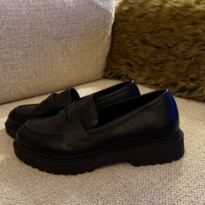 Chunky svarta loafers i skinn - Säljer ett par chunky svarta loafers från Copenhagen Shoes i storlek 38. Skorna har en grov platt sula, rund tå och är tillverkade i slätt skinn. Klassisk design med bred rem över foten, perfekt för dig som gillar stilrena och trendiga skor.