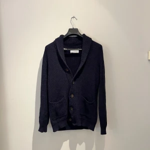 Mörkblå kofta från Selected Homme - Snygg mörkblå stickad kofta från Selected Homme i storlek M. Koftan har grovstickad struktur, bred krage, två framfickor och knappar framtill. Perfekt för lager-på-lager och chill stil. Passar dig som gillar klassiska och stilrena plagg. Hör av er vid frågor eller fler bilder