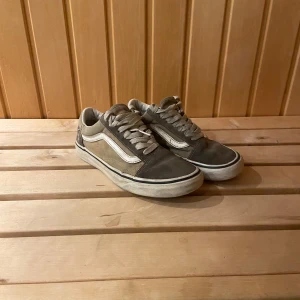 Beige och grå Vans Old Skool sneakers - Säljer ett par klassiska Vans Old Skool sneakers i beige och grått med vit sidorand. Skorna har snörning, platt sula med det ikoniska rutmönstret undertill och rund tå. Ovandelen är i mocka och canvas, vilket ger en skön och cool vibe.
