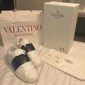 Valentino skor - Stilrena vita sneakers från Valentino Garavani med en bred svart läderdetalj över vristen. Klassisk låg modell med rund tå och vita snören. Snygg och lyxig design i premiumläder, perfekt för dig som gillar exklusiva märken. Skorna är helt nya och kan diskutera pris vid snabb affär!