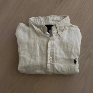 Vit skjorta från Ralph Lauren - Vit skjorta från Ralph Lauren i linne med korta ärmar och klassisk krage. Den har knappar framtill och den ikoniska broderade Polo-loggan i svart på bröstet. Perfekt för en clean och chill look.