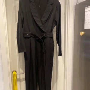 Svart jumpsuit med kavajkrage från NA-KD - Stilren svart jumpsuit från NA-KD med lång ärm, kavajkrage och omlott-design. Midjan markeras med ett bälte med silverdetaljer. Perfekt för dig som gillar en dressad look med edge. Materialet är mjukt och följsamt för en snygg passform.