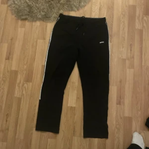 Svarta mjukisbyxor från BOSS XL - Svarta mjukisbyxor från BOSS i storlek XL med vita ränder längs sidorna och snörning i midjan. Byxorna har en enkel och clean design med BOSS-logga på ena benet. Perfekta för chill eller träning. Passar till storlek L