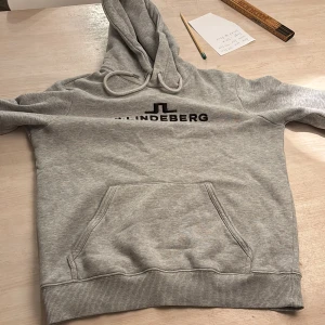 Grå hoodie från J.Lindeberg - Snygg grå hoodie från J.Lindeberg med svart logga på bröstet. Klassisk känguruficka framtill, ribbade muddar och huva med dragsko. Perfekt för chill dagar eller när du vill ha en clean streetstyle.