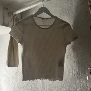 Vit ribbad topp från ONLY - Säljer en vit ribbad topp från ONLY i storlek M. Toppen har korta ärmar, rund halsringning och ett diskret randigt mönster. Den är croppad med vågiga kanter vid ärmslut och nederkant, vilket ger en söt och trendig vibe. Den sitter fint på kroppen och passar mig som har storlek S/XS