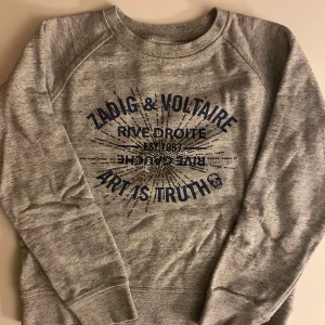 Grå sweatshirt Zadig & Voltaire - Snygg grå sweatshirt från Zadig & Voltaire med blått tryck framtill där det står 'Art is Truth'. Tröjan har rund halsringning, långa ärmar och mjuk bomullskänsla. Perfekt för en avslappnad och cool stil. BARN STORLEK ÅR 10 ÅR