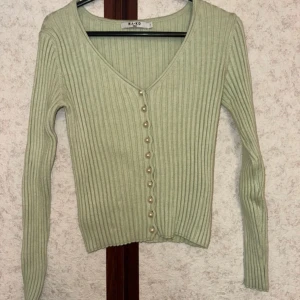 Ljusgrön ribbad kofta från NA-KD - Superfin ljusgrön ribbad kofta från NA-KD med v-ringning och pärlknappar framtill. Långärmad och figurnära passform, perfekt att styla med jeans eller kjol. Materialet är mjukt och stretchigt för en bekväm känsla.