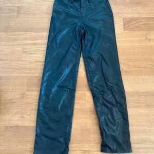 Svarta skinnbyxor från H&M Divided - Snygga svarta byxor  från H&M Divided. Byxorna har raka ben, hög midja och dragkedja framtill med knapp. Perfekta för dig som vill ha en edgy look med lite glansig finish.Onavänd!