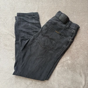 Svarta Nudie Jeans - Säljer ett par svarta jeans från Nudie Jeans i storlek 33/32. Klassisk femficksmodell med silverfärgade knappar och svarta sömmar på bakfickan. Jeansen har rak passform och är tillverkade i mjukt bomullstyg. Perfekta för en clean och stilren look.