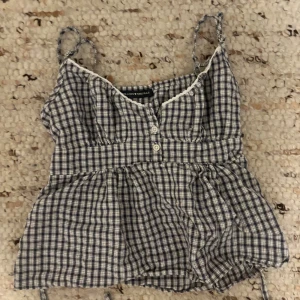 Rutig blå linnetopp från Brandy Melville - Supersöt rutig linnetopp från Brandy Melville i blått och vitt. Toppen har smala axelband, knappar framtill och en söt spetskant vid halsen. Den har en knytning i ryggen och är lätt rynkad för en chill och feminin vibe. Perfekt för varma dagar!