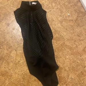 Svart asymmetrisk klänning Rare London - Svart body  från Rare London med hög hals. Bodyn är ärmlös och har ett snyggt strukturerat mönster framtill. Stängs med dragkedja i ryggen. Perfekt för dig som ha en fest outfit