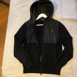 Svart hybrid jacka från J.Lindeberg - Svart vindjacka från J.Lindeberg med huva och hel dragkedja. De är en mestadels stickad jacka med polyester där uppe. Passar perfekt hela året. Finns lite färgstänk längst ner på höger ärm men inget man märker. NYPRIS runt 3000 köpt i butik Stockholm 