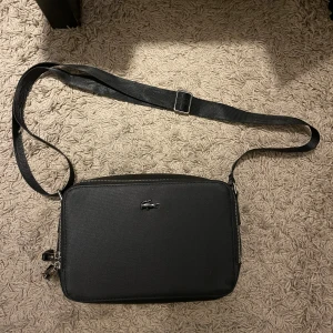 Svart axelväska från Lacoste - Snygg svart axelväska från Lacoste med klassisk krokodillogo framtill. Väskan har en justerbar axelrem med diskret logga, två dragkedjor och stilren, rektangulär form. Materialet är slitstark polyester som ger en clean och modern look.