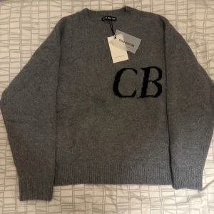 Grå stickad tröja från Cole Buxton - Snygg grå stickad tröja från Cole Buxton med svarta CB-bokstäver på bröstet. Tröjan har rund halsringning, långa ärmar och ribbade muddar vid ärmslut och nederkant. Perfekt för dig som gillar stilrena och bekväma plagg.