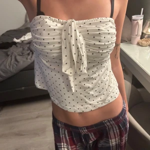 Vit prickig tubtopp med knyt - Säljer en söt vit tubtopp med svarta prickar och knyt framtill. Toppen är ärmlös och har en bandeau-formad överdel som sitter uppe utan axelband. Perfekt för varma dagar och ger en fräsch och trendig look.