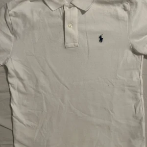 Ralph Lauren Pike - Vit pikétröja från Polo Ralph Lauren i slim fit-modell. Klassisk krage, kort ärm och knäppning framtill med två knappar. Ikonisk marinblå broderad logga på bröstet. Tillverkad i mjuk bomull som känns skön mot huden.
