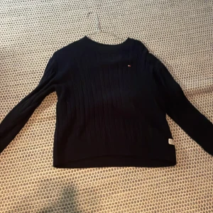 Mörkblå stickad tröja Tommy Hilfiger - Snygg mörkblå stickad tröja från Tommy Hilfiger med diskret logga på bröstet. Tröjan har lång ärm, rund halsringning och ribbade detaljer. Perfekt för dig som gillar klassisk och stilren look.