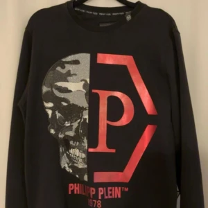 Svart Philipp Plein sweatshirt med tryck - Svart sweatshirt från Philipp Plein med stort rött logotryck och cool dödskalle i silver och camouflage på framsidan. Tröjan har rund halsringning och långa ärmar. Perfekt statement piece för dig som gillar streetstyle.