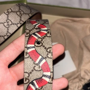 Gucci beige bälte med ormprint - Säljer ett Gucci bälte i beige med klassiskt GG-mönster och en cool röd, vit och svart orm som slingrar sig över hela bältet. Bältet är tillverkat i skinn och har en bred, platt form som sticker ut. Perfekt för dig som vill ha något unikt och trendigt från Gucci. Äkta bälte
