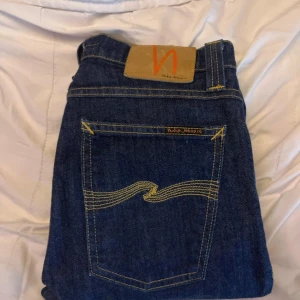 Blå jeans från Nudie Jeans Co W30 L34 - Säger ett par helt oanvända snygga blå jeans från Nudie Jeans Co med klassisk gul kontrastsöm och ikonisk broderad bakficka. Modellen har raka ben och normal passform. Tillverkade i slitstarkt bomullstyg, perfekta för en avslappnad och stilren look, hör eh dig vid frågor!