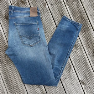 Replay Anbass blå jeans herr 33/32 - Säljer ett par Replay Anbass jeans i klassisk blå tvätt med snygga slitningar. Modellen har raka ben, fem fickor och detaljerade sömmar på bakfickorna. Jeansen är tillverkade i bomull och har en normal passform. Perfekta för dig som gillar stilrena och bekväma jeans. Kom gärna med frågor/offer!🙌