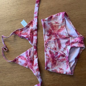 Rosa bikini med palmmönster - Snygg bikini från Kappahl i rosa och ljusrosa toner med tropiskt palmmönster. Överdelen har trekantsform och knyt i nacken, medan underdelen är klassisk med hög midja. Perfekt för strandhäng och poolparty!