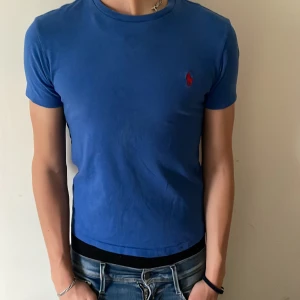 Blå t-shirt från Ralph Lauren - Snygg blå t-shirt från Ralph Lauren med klassisk röd logga broderad på bröstet. Modellen är slim och har rund hals samt korta ärmar. Materialet är mjuk bomull som sitter skönt mot kroppen. Perfekt för dig som gillar stilrena och tidlösa plagg. Storlek xs skick 9/10 använd bara några få gånger säljer då jag har växt ur den. Pris går att diskutera. 