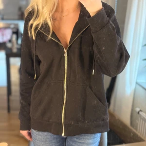 Svart hoodie med dragkedja i storlek L - Svart hoodie med huva och snörning, hel dragkedja i guld och två fickor framtill. Tillverkad i mjuk bomullsmix, perfekt för chill dagar. Klassisk passform och långa ärmar.